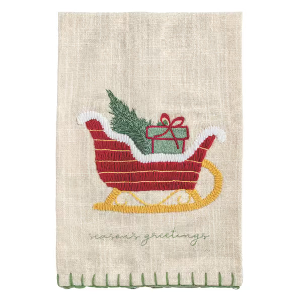 A beige linen tea towel with a festive embroidered design of a red sleigh carrying a green pine tree and a red gift box, with 'seasons greetings' stitched below.