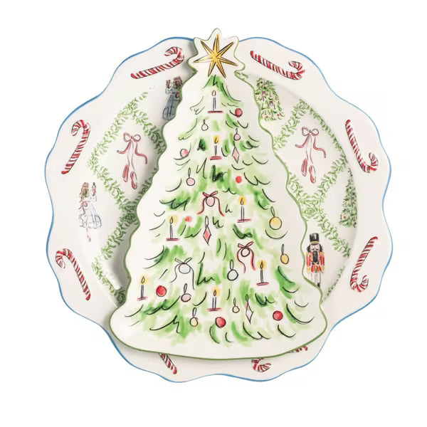A decorative ceramic Christmas tree-shaped plate or dish placed on top of a scalloped round plate with holiday illustrations including candy canes, green garlands, and a nutcracker, shown on a white background.