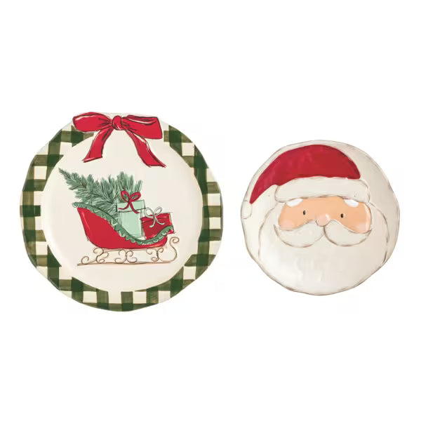 Two Christmas-themed ceramic plates on a white background. One round plate features a green and white checkered border, a red bow, and an illustration of a sleigh carrying green pine branches and red gifts.