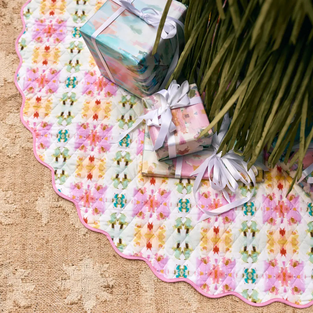 A scalloped tree skirt with a colorful floral pattern and pink edge, styled on a burlap surface with wrapped gift boxes and green palm fronds.
