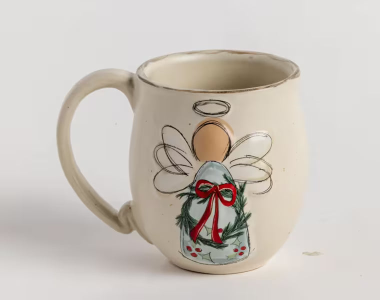 An off-white ceramic mug featuring a painted Christmas angel design with green holly leaves, a red bow, and a gold halo, set against a plain light background.