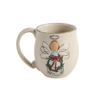 An off-white ceramic mug featuring a hand-painted Christmas angel design with a green wreath, red bow, and gold details, set against a plain white background.