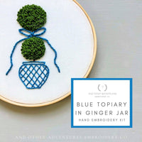 Embroidery hoop with blue topiary design and text about embroidery kit on a light gray background