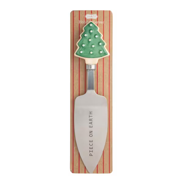 A Christmas-themed serving spoon with a green tree-shaped handle and a silver metal blade engraved with 'PIECE ON EARTH', mounted on a striped wooden display card.