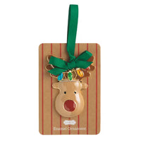 A Christmas enamel ornament shaped like a reindeer head with gold-tone antlers, a green ribbon loop, and colorful jewel-like lights, attached to a brown striped backing card labeled Enamel Ornament.