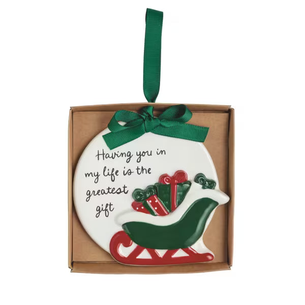 A round white Christmas ornament in a kraft gift box, featuring a green hanging ribbon, a green bow, and a red sleigh carrying green and red presents. The ornament has the phrase 'Having you in my life is the greatest gift' written in black script.