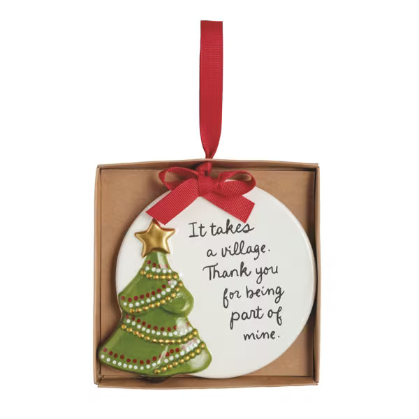 A ceramic Christmas ornament in a square kraft gift box, featuring a white round ornament with a green decorated Christmas tree, a gold star, and the handwritten text 'It takes a village. Thank you for being part of mine.