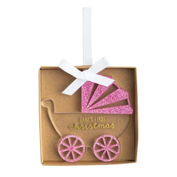 A baby shower ornament shaped like a stroller, featuring a pink glitter canopy and two matching pink glitter wheels, presented in a brown kraft gift box with a white ribbon and gold 'Baby's First Christmas' text.