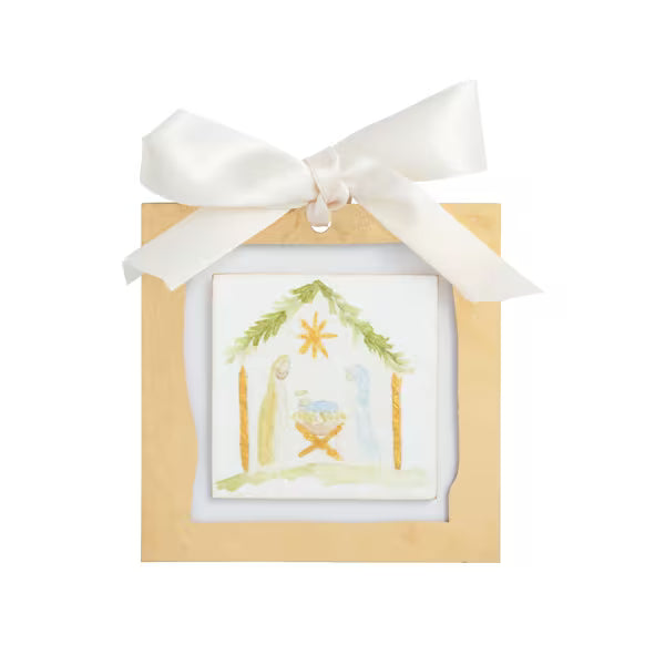 A small square nativity art print displayed inside a light wood frame, tied with a white satin ribbon at the top. The print shows a simple nativity scene with a stable, star, and figures in soft pastel colors.