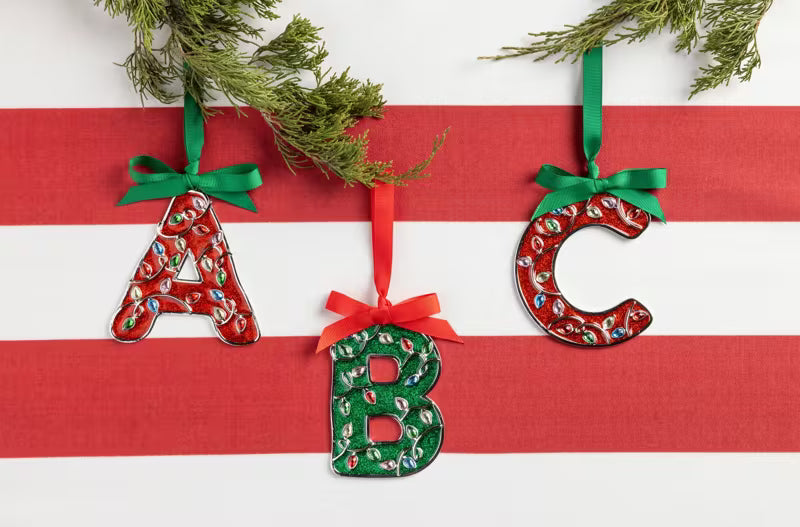Three decorative hanging letters A, B, and C made from red and green holiday-patterned fabric, suspended from green ribbon bows and evergreen branches against a red-and-white striped background.