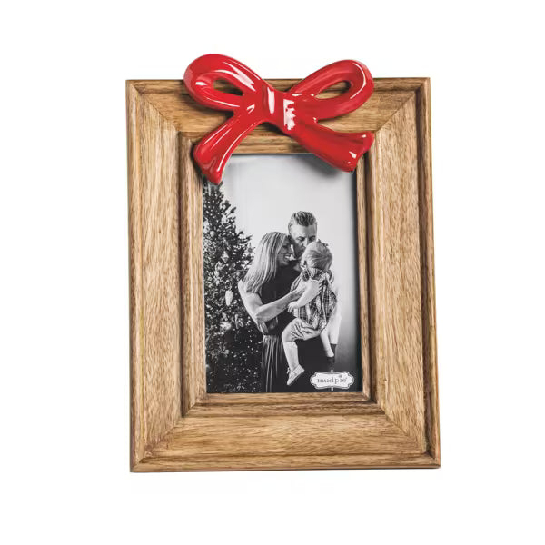 A square wooden picture frame with a natural oak-like finish, decorated with a glossy red bow at the top. The frame holds a black-and-white photo of a family, and a small white nameplate is visible in the lower right corner of the insert.