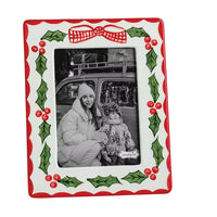 A square white ceramic Christmas-themed picture frame with a red border, green holly leaves, red berries, and a red-and-white checkered bow at the top. The frame holds a black-and-white photo of a woman and child in front of a vehicle.