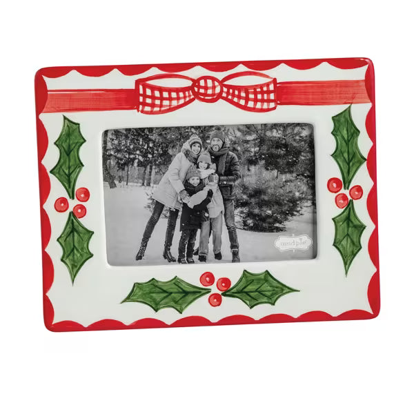 A rectangular ceramic picture frame with a festive Christmas design, featuring a red and white scalloped border, a red bow at the top, green holly leaves, and red berries. The center is blanked out with a placeholder family photo.