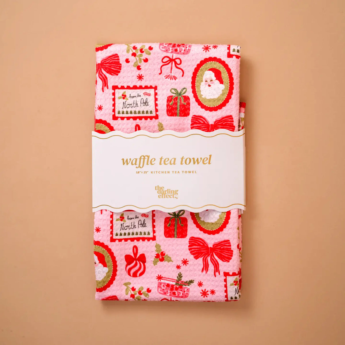 A pink waffle tea towel with a festive Christmas print, wrapped in a white paper band that reads 'Vintage Sheets'. The towel is displayed flat against a solid beige background.