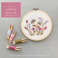 Embroidery hoop with floral design and embroidery thread clips on a gray background, featuring 'Jardín Mauve' embroidery kit by And Other Adventures Embroidery Co.