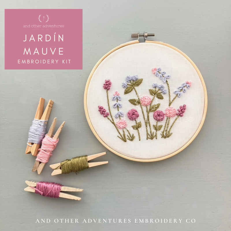 Embroidery hoop with floral design and embroidery thread clips on a gray background, featuring 'Jardín Mauve' embroidery kit by And Other Adventures Embroidery Co.