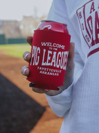 Red can cooler with 'Welcome to the PIC LEAGUE! Fayetteville Arkansas' text held by a person on a baseball field.