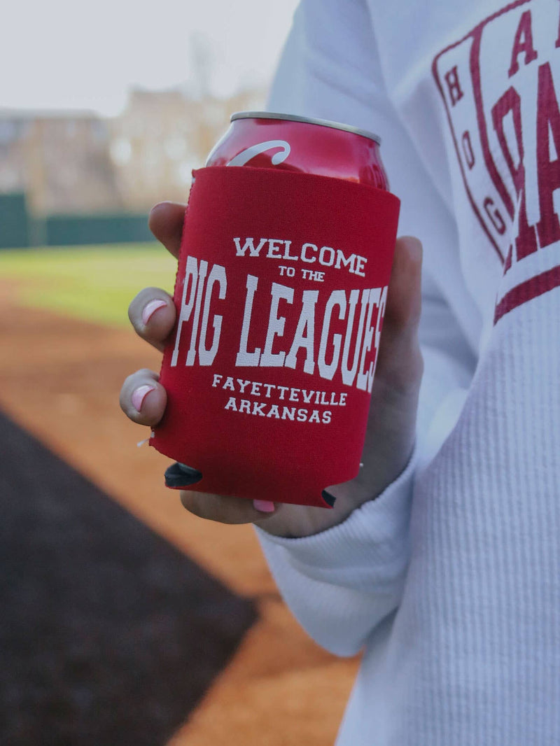 Red can cooler with 'Welcome to the PIC LEAGUE! Fayetteville Arkansas' text held by a person on a baseball field.