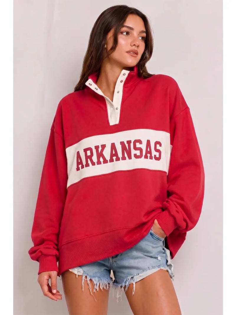 A woman wearing a red long-sleeve quarter-zip sweatshirt with a white chest stripe featuring the word 'ARKANSAS' in red, paired with light-wash denim shorts, against a plain light gray background.