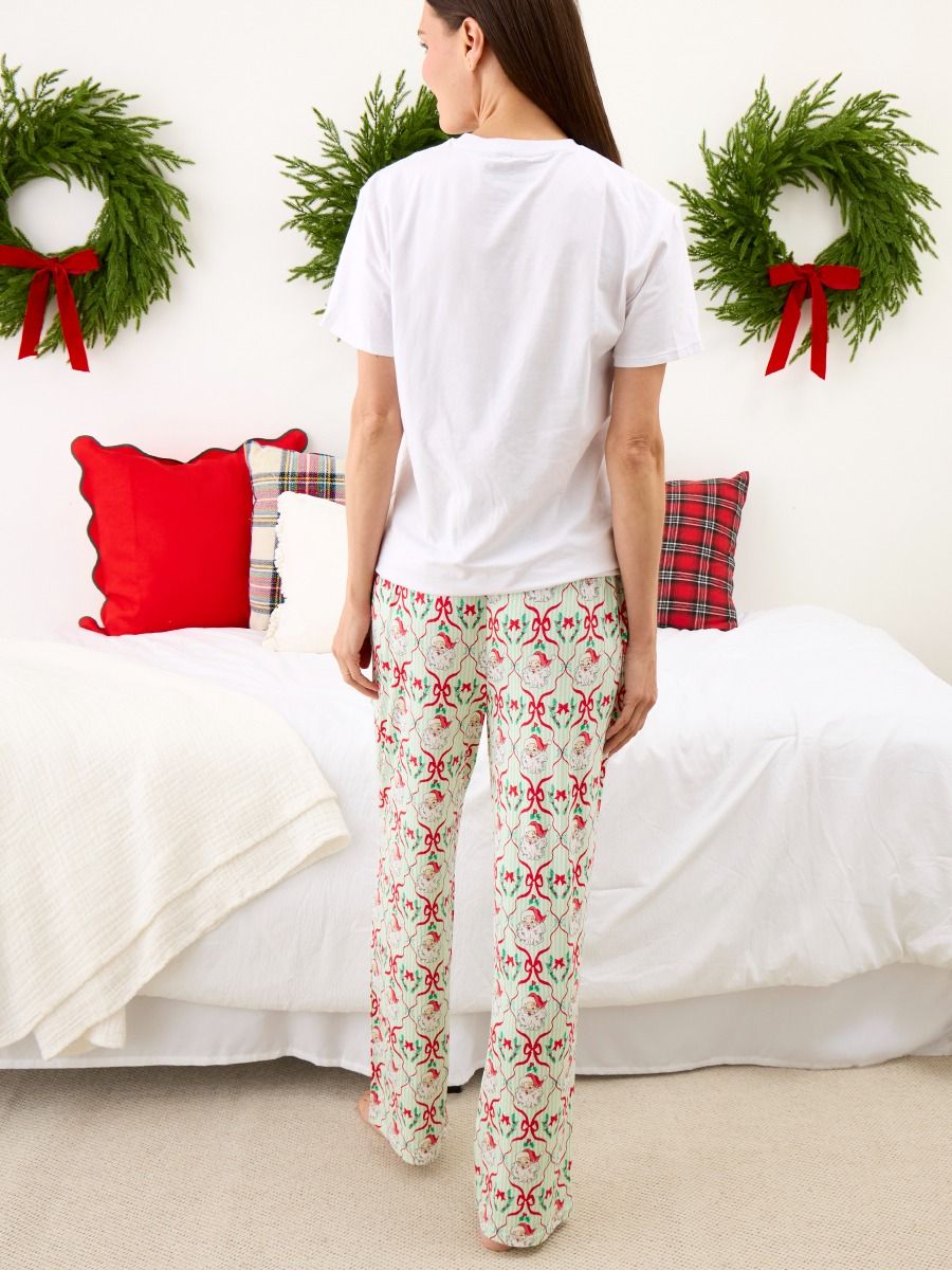 A woman stands with her back to the camera in a bedroom, wearing a white t-shirt and holiday-themed patterned pajama pants. The pants feature a light cream background with a repeating red and green Christmas ornament motif.