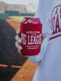 Person holding a red can cooler with 'Welcome to the Pig League' text on a baseball field.