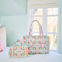 Floral patterned duffel bag and travel case on a bed with a window in the background.