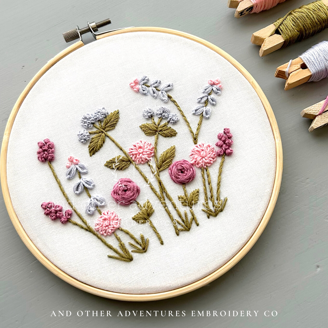 Embroidery hoop with floral embroidery on a gray surface, featuring 'And Other Adventures Embroidery Co'.