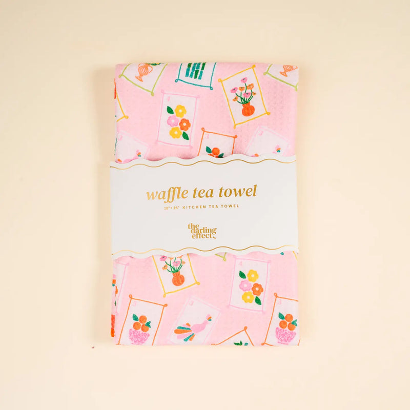 A folded pink waffle tea towel with a floral and teacup pattern, wrapped in a white paper band that reads 'waffle tea towel' and 'The Time Traveler', set against a solid beige background.
