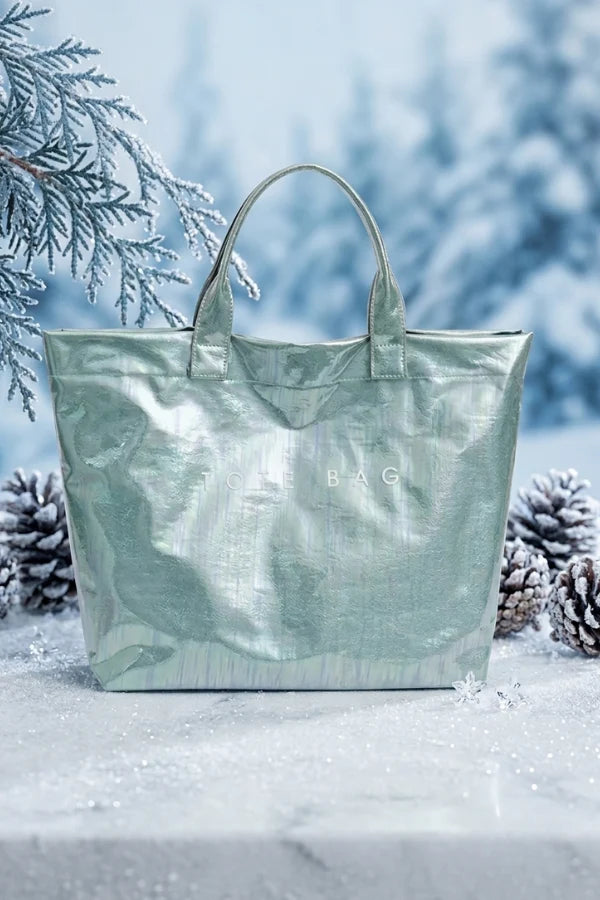 Light green tote bag with 'BAG' branding on a snowy surface with pine branches and cones.