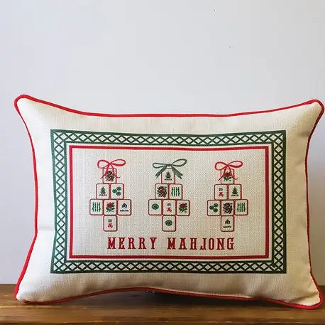 A rectangular decorative throw pillow with a Mahjong-themed cross-stitch style print. The pillow has a cream fabric base with red piping, a green and red border, and three decorated tiles with the text 'MERRY MAHJONG' underneath, resting on a wooden surface against a plain white wall.