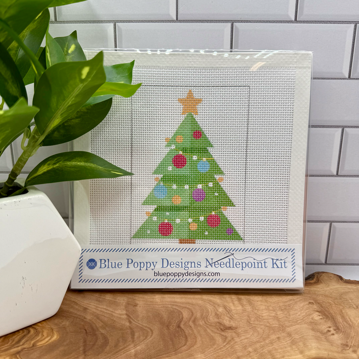 Cross-stitch kit with a Christmas tree design on a wooden surface next to a plant.