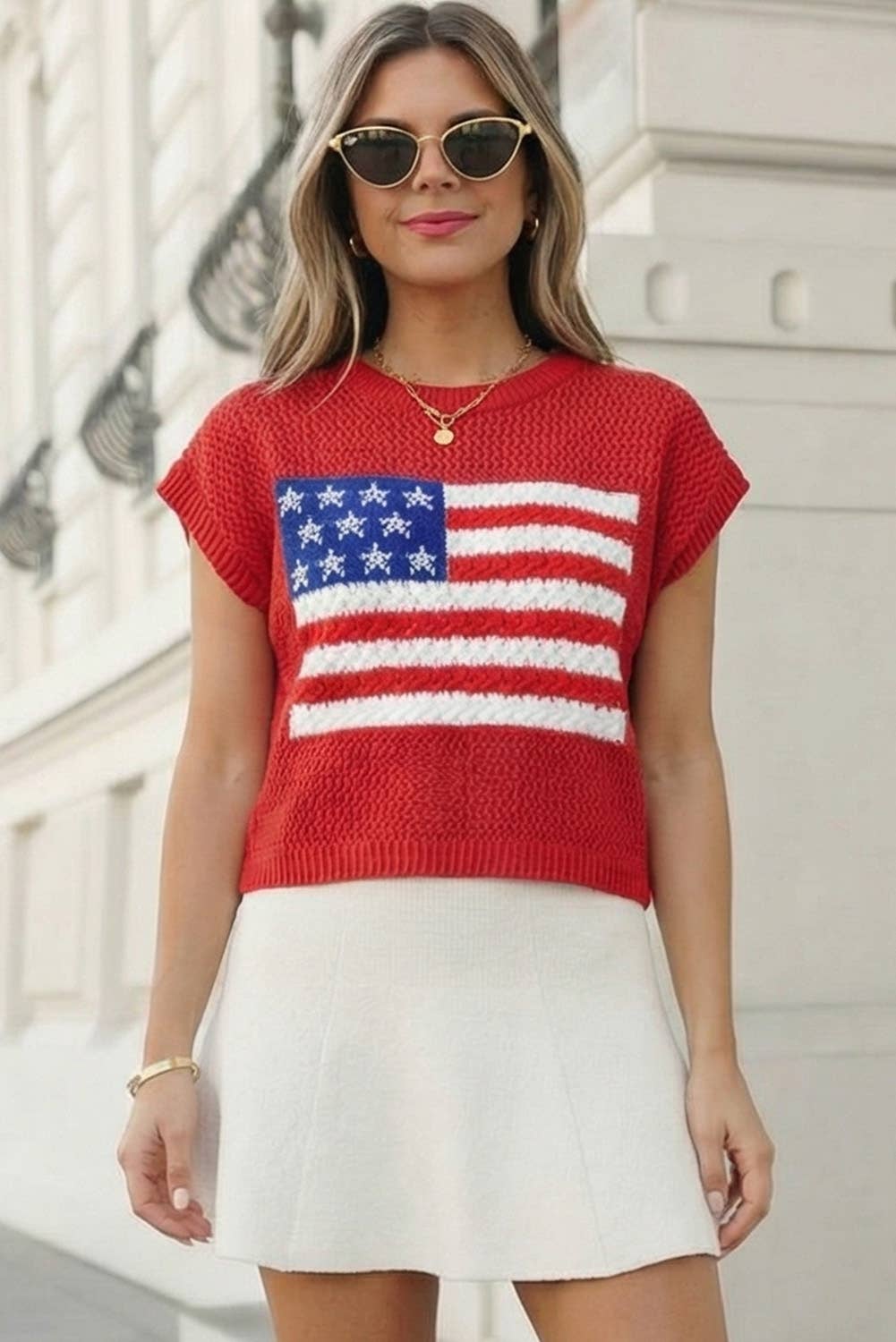 Woman wearing a red sweater with an American flag design, sunglasses, and a white skirt.