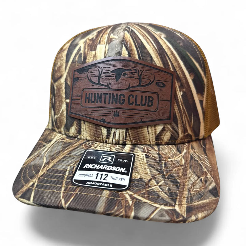A Richardson 112 trucker hat in a brown and beige marsh grass camouflage pattern, featuring a brown leather 'HUNTING CLUB' patch on the front and a Richardson brand sticker on the visor.