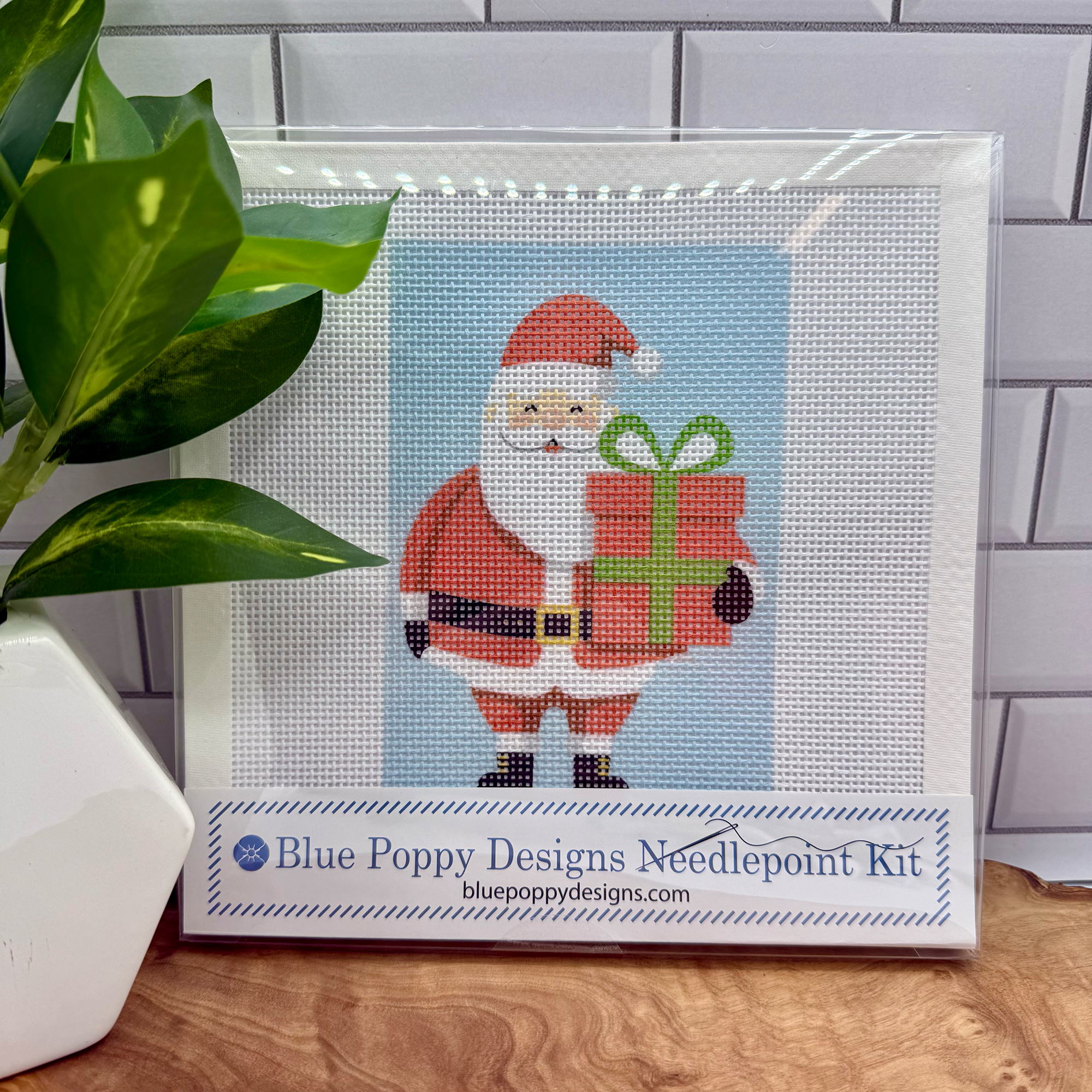 Cross-stitch kit with Santa Claus design by Blue Poppy Designs on a wooden surface with a plant.