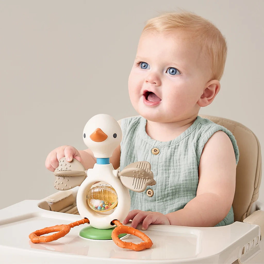 A baby sitting in a high chair holds a duck-shaped sensory toy with orange rings and textured panels on a light beige background.