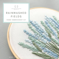 Hand embroidery kit with embroidered plants on a gray background, featuring the brand 'Rainwashed Fields'.