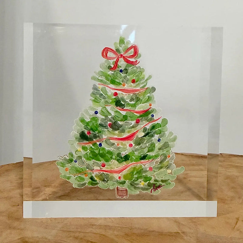 A square clear acrylic block featuring a hand-painted watercolor Christmas tree with green foliage, red ribbons, and ornaments, displayed on a wooden surface against a white background.
