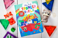 Colorful children's book titled 'My First Toddler Bible' surrounded by toys on a wooden surface.