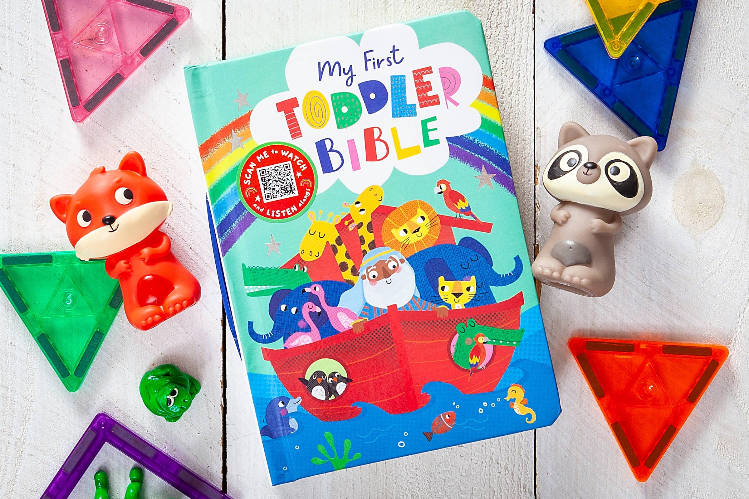 Colorful children's book titled 'My First Toddler Bible' surrounded by toys on a wooden surface.