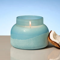 Candle in a blue glass jar with a lighted wick, placed on a reflective surface with a blurred background.