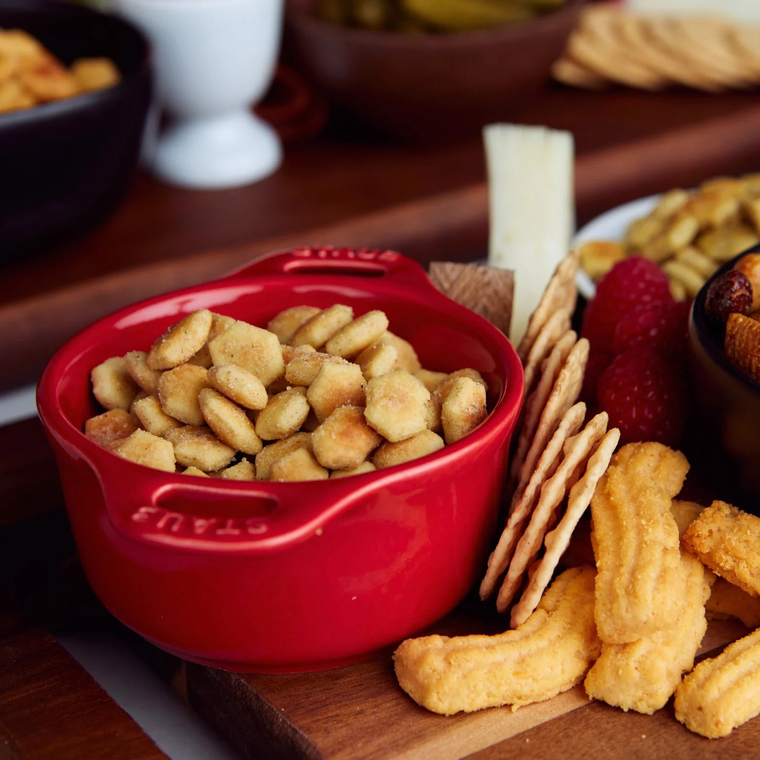 Red bowl of oyster crackers on a wooden board with crackers and other snacks.