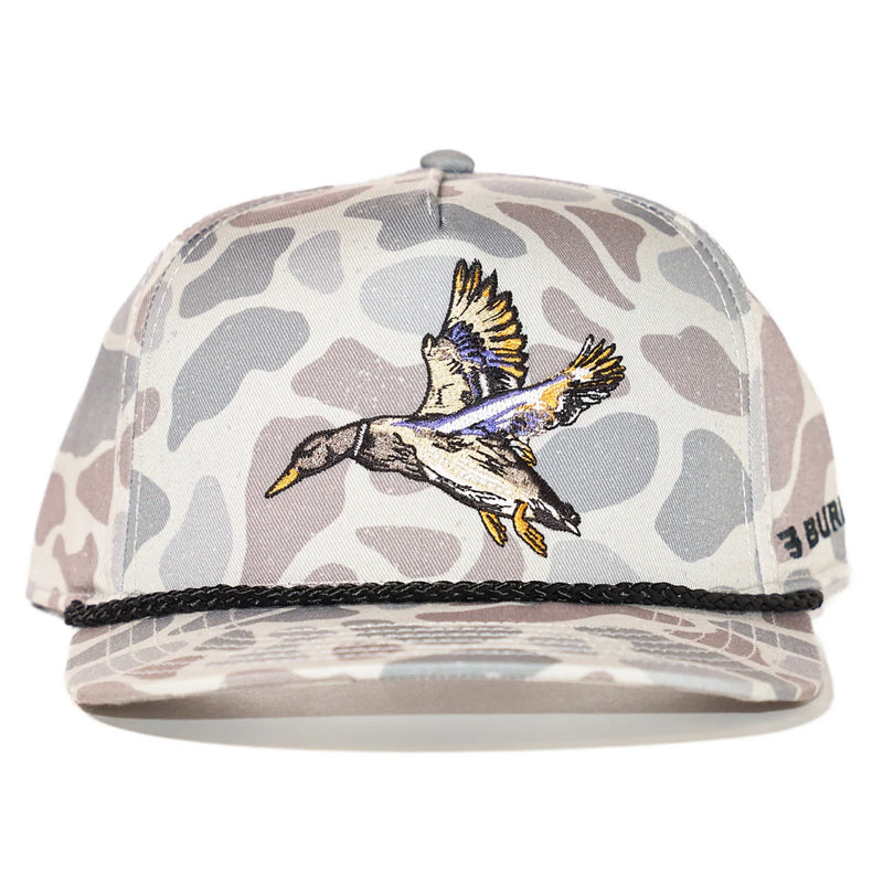 A camouflage trucker cap featuring a flying mallard duck embroidered on the front panel, a black braided rope across the brim, and a small black brand patch on the side.