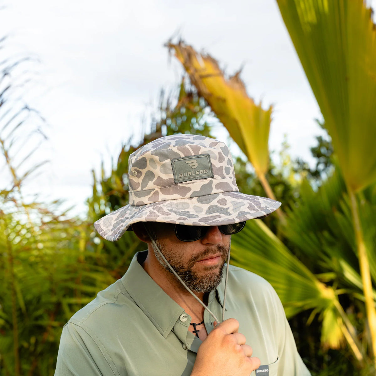 Man wearing a camouflage bucket hat with a brand logo, standing in a tropical setting.