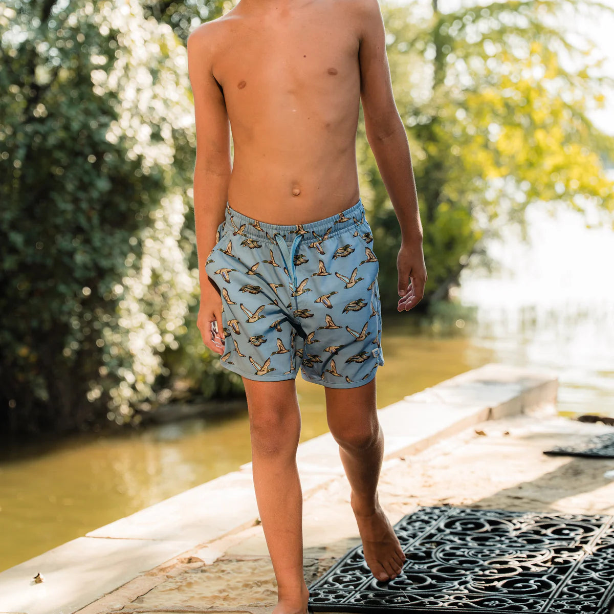 Person wearing blue swim shorts with a nature pattern by a body of water.