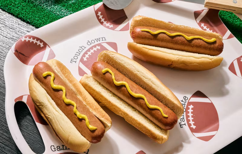 Three hot dogs in buns with mustard on top, arranged on a white football-themed serving tray with printed football graphics and the words 'touch down' and 'Gamecocks'.