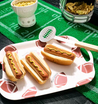 A white rectangular serving tray with football-themed graphics and handles, holding three hot dogs in buns with mustard. A metal hot dog turner with a green stripe pattern rests on the tray.