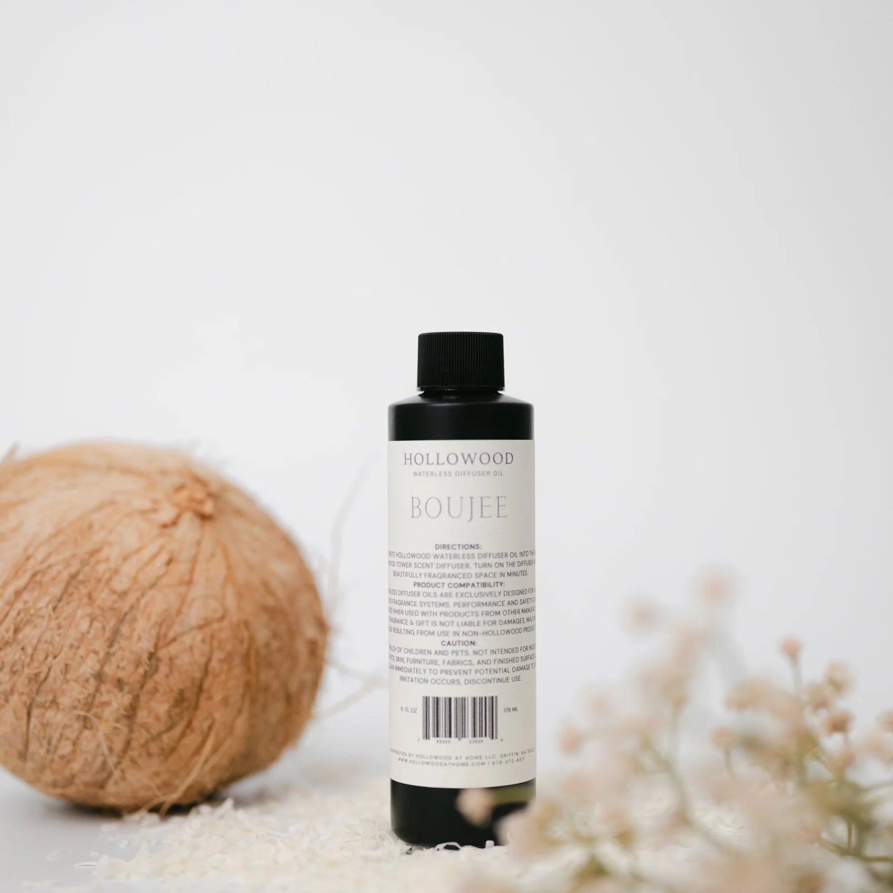 Bottle of Hollywood Boujee product with a coconut and flowers on a light background