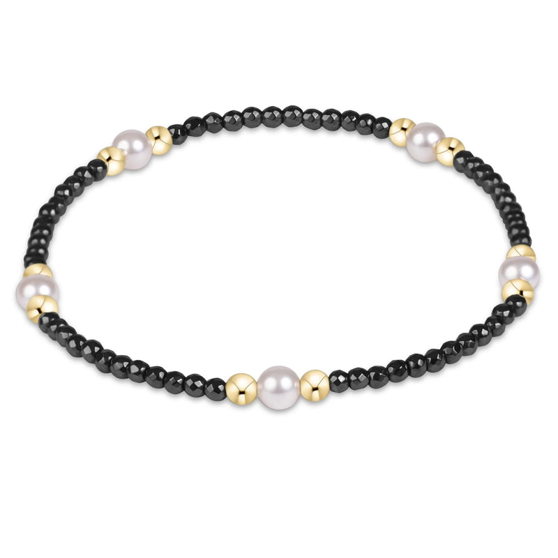 A beaded stretch bracelet featuring small black faceted beads, larger polished black beads, white pearls, and shiny gold spacer beads, arranged in a circular shape on a plain white background.