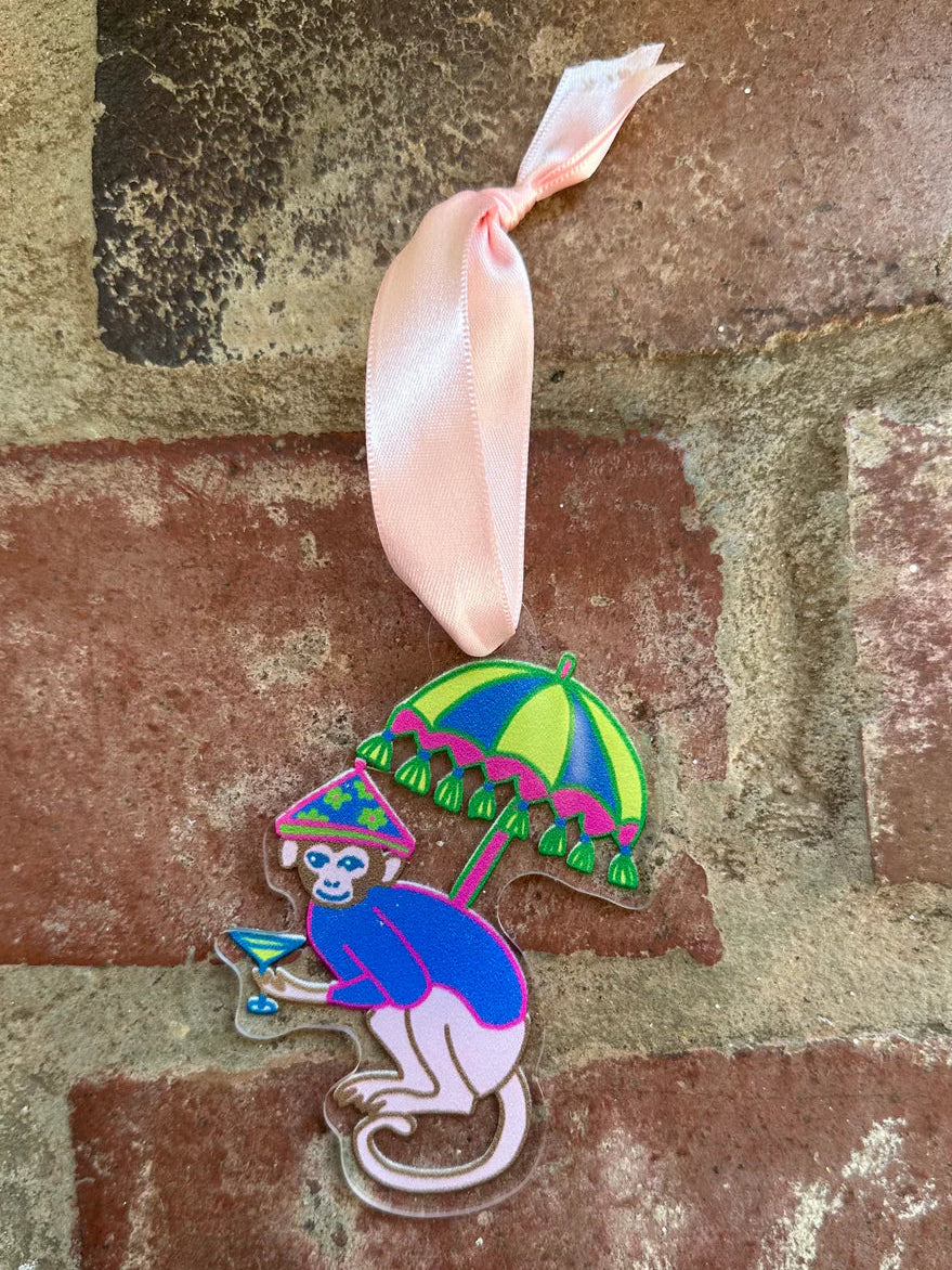 A colorful enamel pin shaped like a monkey sitting under a striped green and pink umbrella, hanging from a light pink ribbon, displayed on a rustic red brick background.
