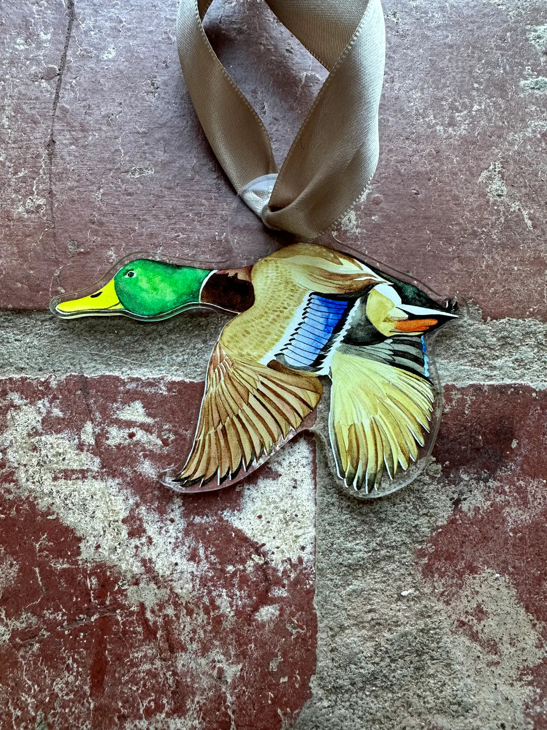A handcrafted wooden duck ornament with a tan ribbon hanger, resting on a weathered red-brown brick surface. The duck is painted with detailed naturalistic colors including a green head, yellow beak, brown and gold wings, and blue accents.